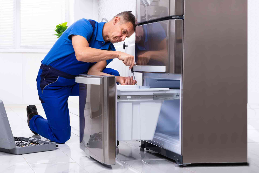 Appliance repair technician kneeling on one knee while using a screwdriver to remove or replace a bottom freezer drawer component on a stainless steel refrigerator, with tools and an open toolkit on the floor in a bright modern kitchen.